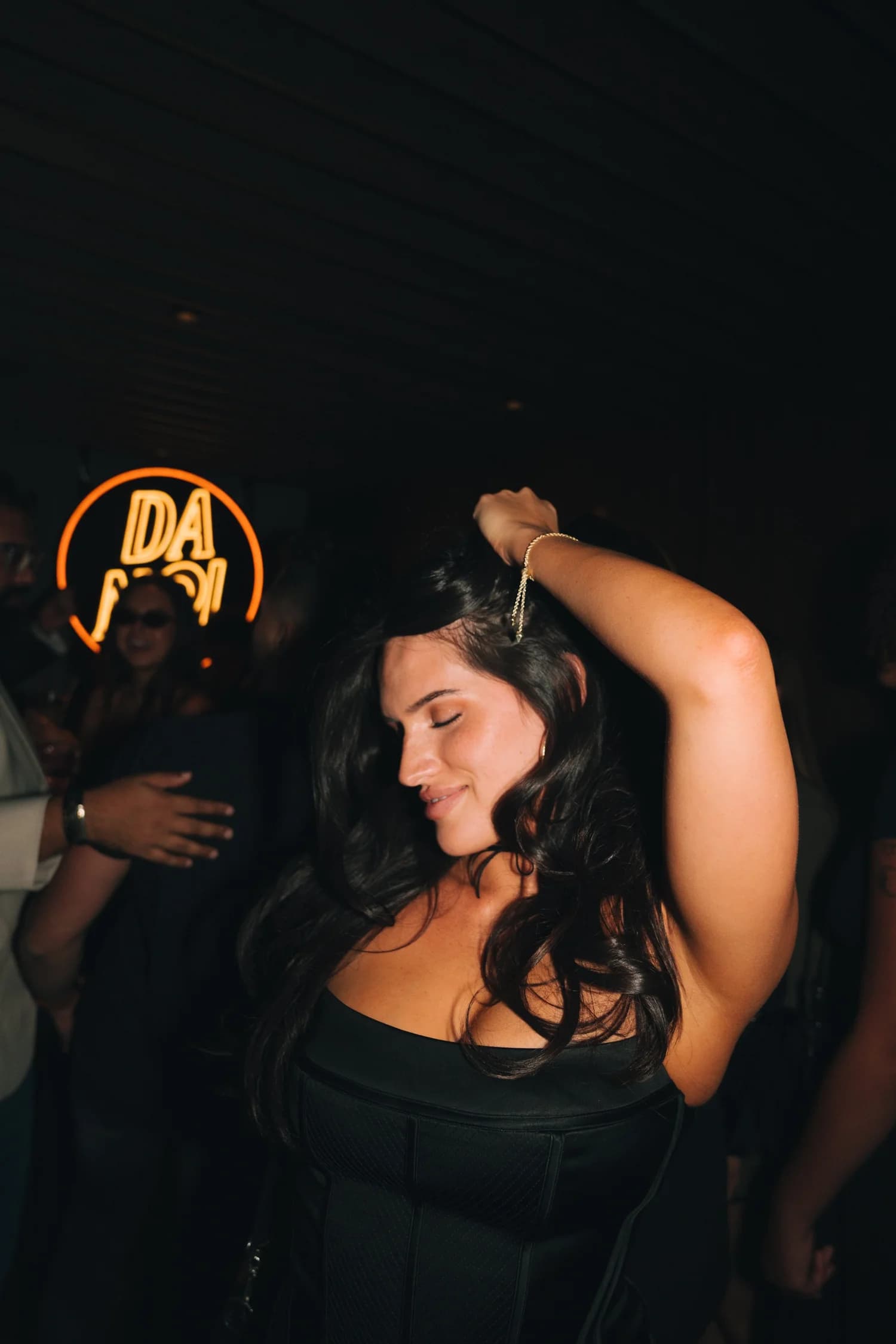 A woman dancing with one arm up under the orange neon Da Noi sign