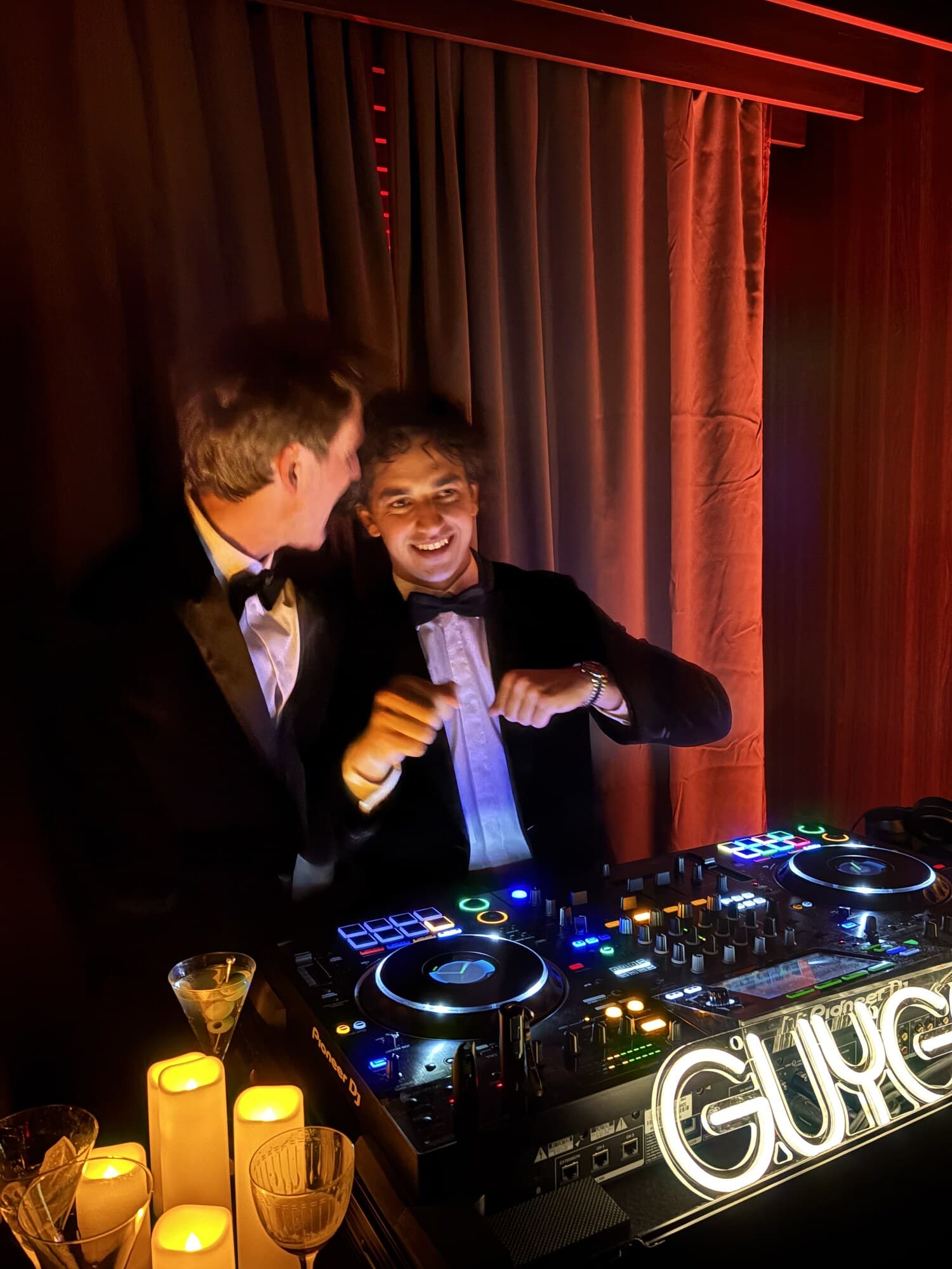 Two DJs in tuxedos laughing together at the decks by candlelight on New Year's Eve